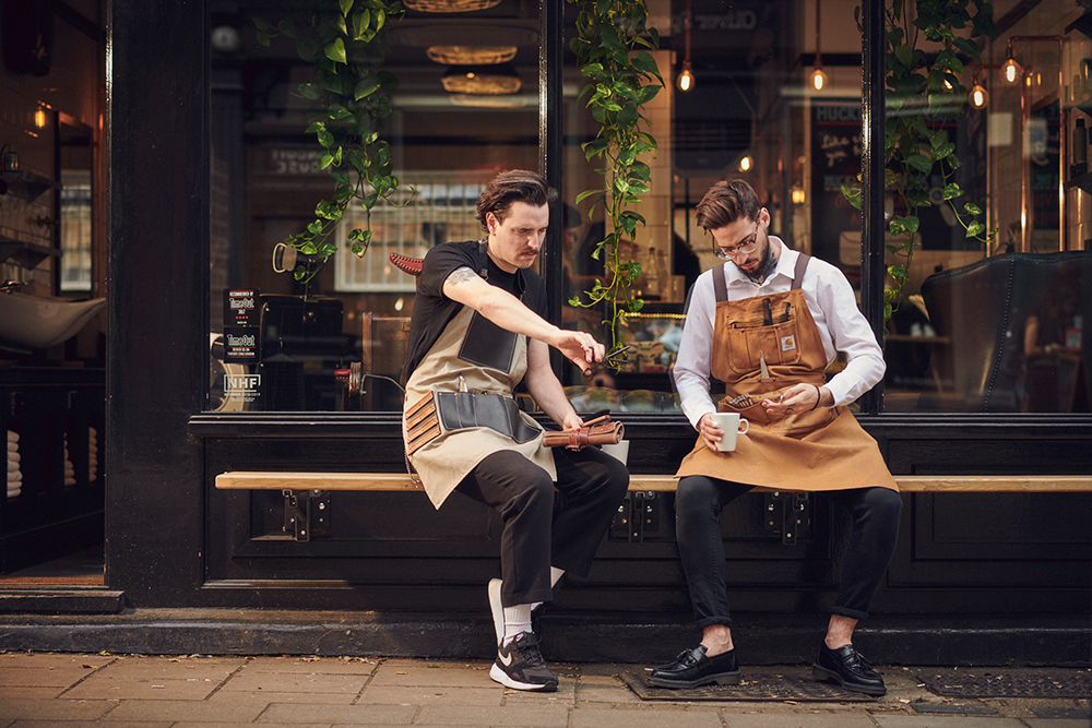 two barbers holding a tool roll and coffee outside a barbershop